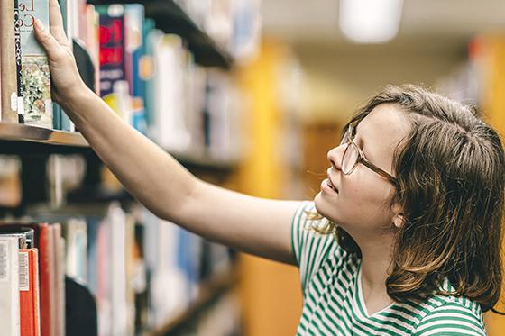 图为一名身穿绿白条纹t恤的波胆网站女学生在珍妮·梅隆·金图书馆的书架上伸手拿书. 