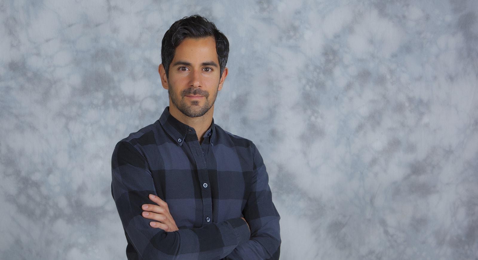 Assistant Professor of Exercise Science at Chatham University, Andres Carrillo, poses with arms crossed in front of grey background. 