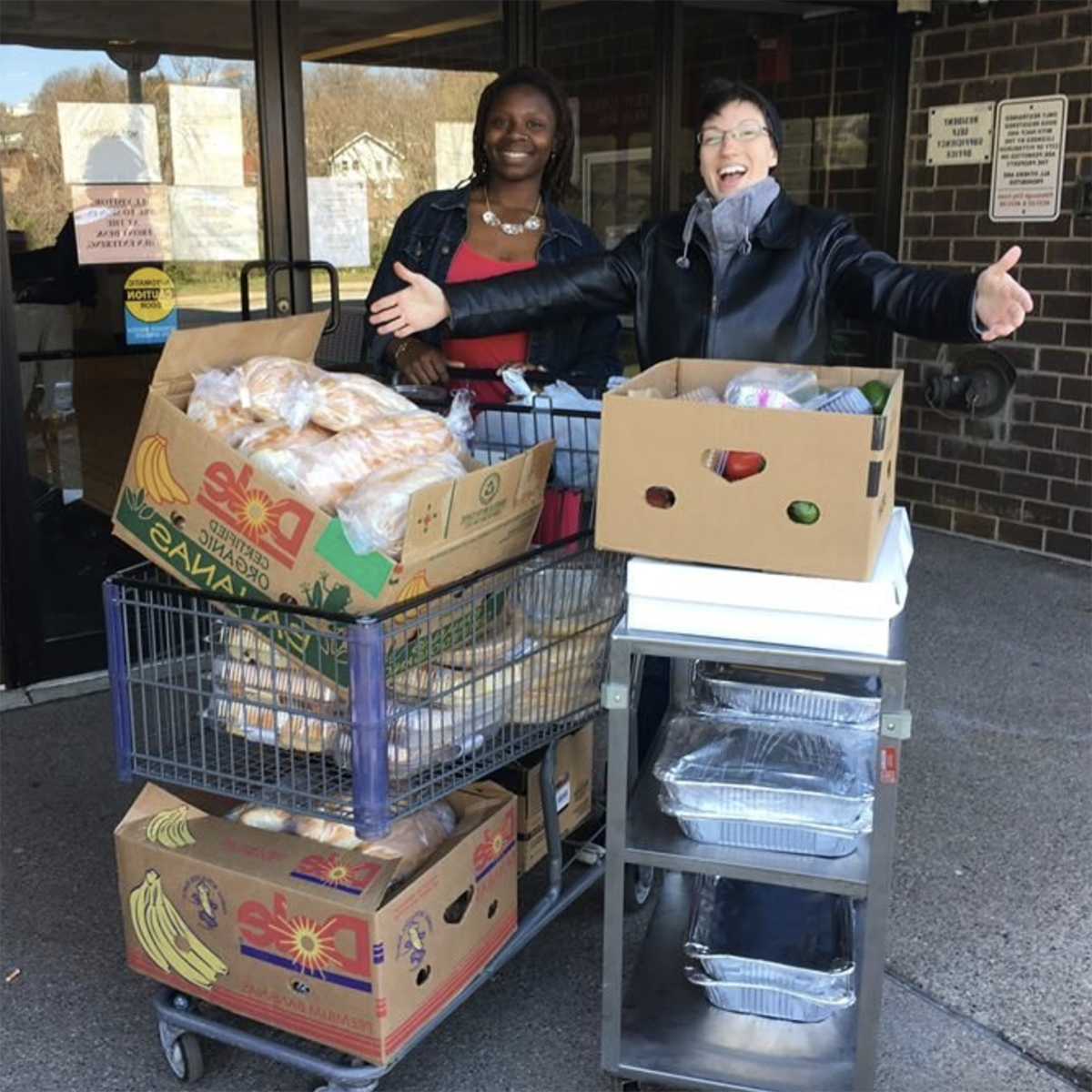 Photo of two people smiling and posing for a photo in front of carts full of food, in front of a grocery store entrance